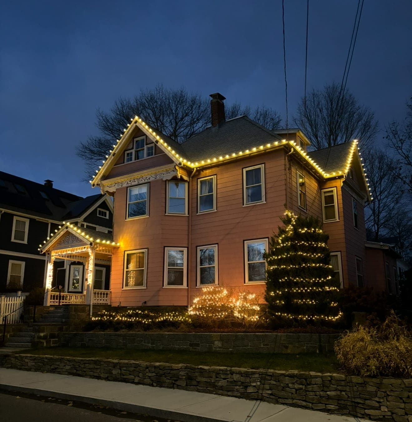 Victorian home with warm-white roofline lights, lit porch gable, lit evergreen and lit shrubs behind a stone wall