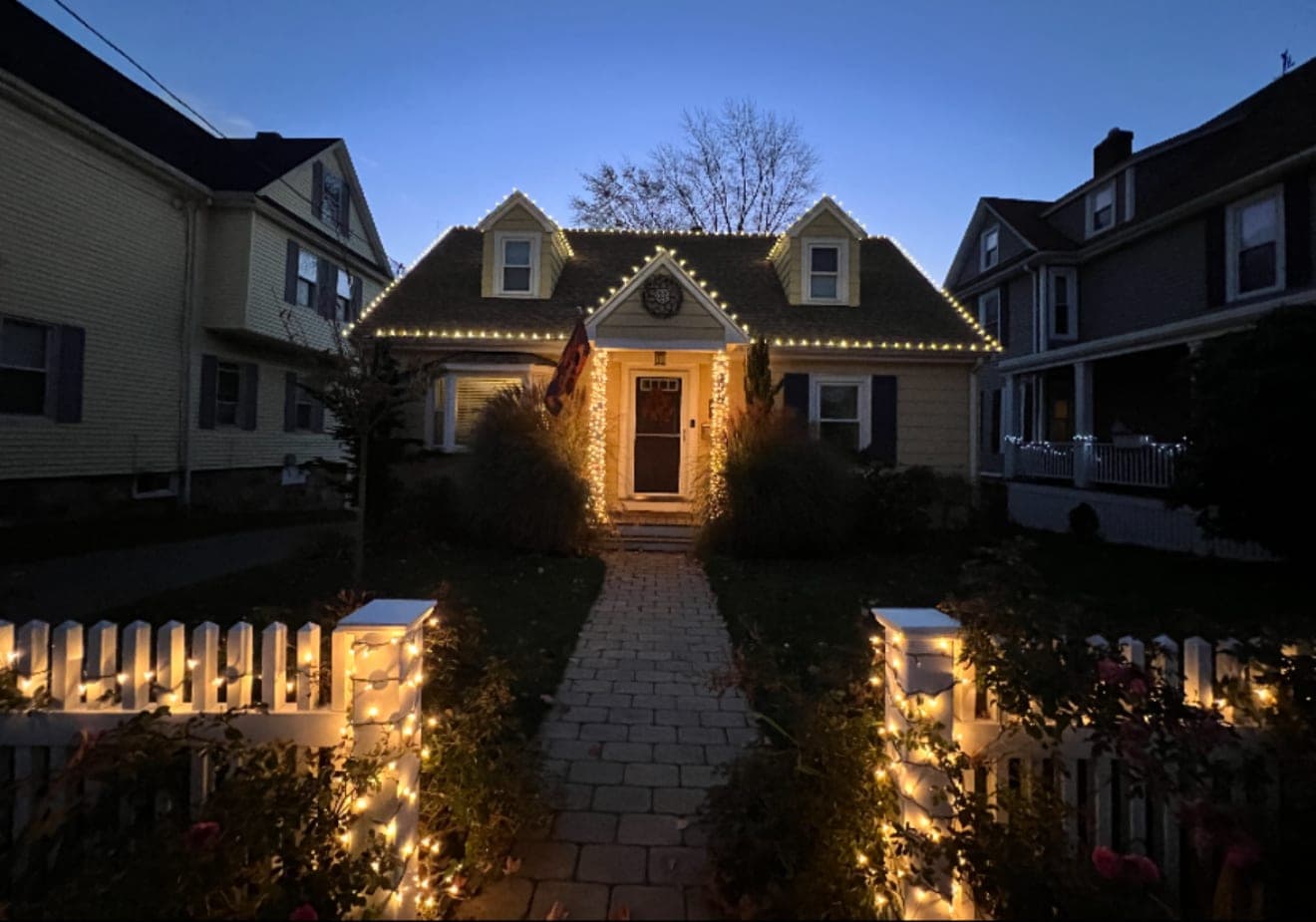 Cape with two dormers, warm-white roofline lights, lit garland at the door and lit fence posts along the walkway