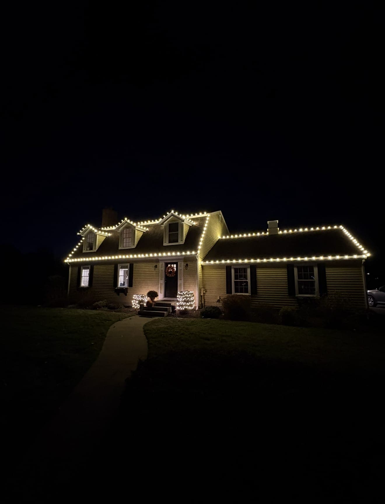 Dark night view of a cape with three dormers, long warm-white roofline and two lit shrubs at the entry