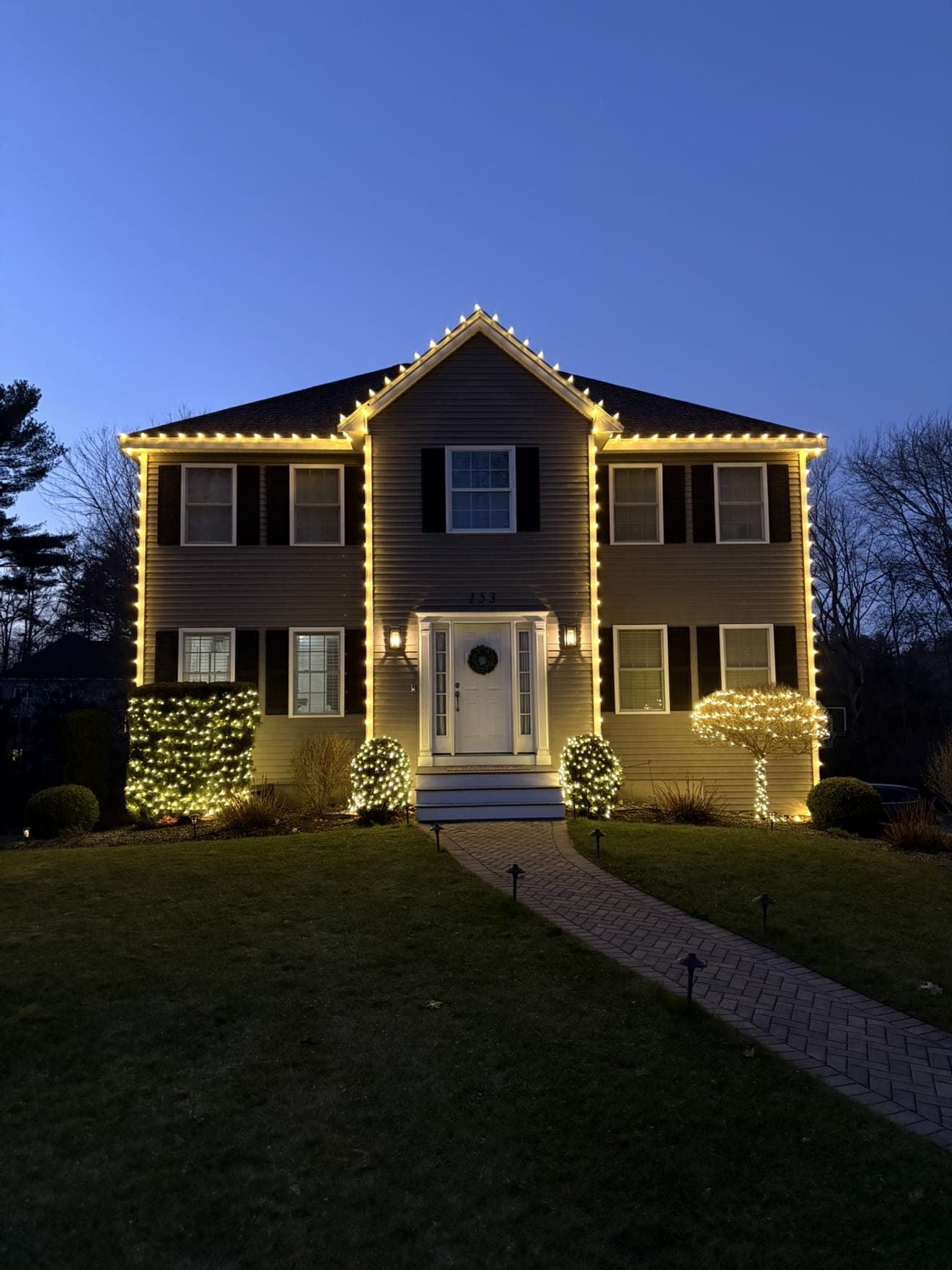 Two-story colonial at blue hour outlined in warm-white roofline lights with lit shrubs and a wreath on the door