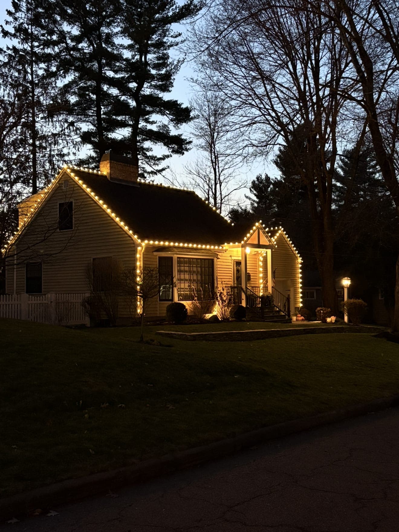 Cape at dusk with warm-white roofline lights and a lit entry gable beside a lamppost